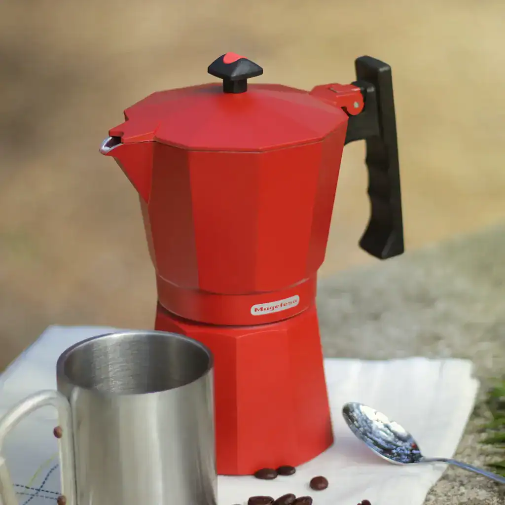 Cafetera moka Magefesa de aluminio roja con taza metálica y granos de café sobre mesa de piedra al aire libre