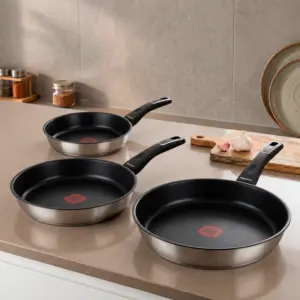 Three Magefesa Onix Red stainless steel fry pans of different sizes arranged on a kitchen countertop with a wooden cutting board and garlic.